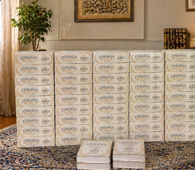 May include: A large stack of beige boxes labeled "ETHIOPIAN BIBLE" in gold lettering. The boxes are neatly arranged, with two open boxes in the foreground. The background includes a framed artwork and a small potted plant.