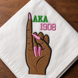 May include: A white fabric napkin featuring an embroidered design of a brown hand with pink manicured nails held in an upward pointing gesture. The text AKA is embroidered in green above the number 1908, which is embroidered in pink thread.