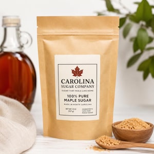 May include: A brown paper bag of Carolina Sugar Company 100% Pure Maple Sugar, with a red maple leaf logo. The bag is labeled "Made in North Carolina" and contains 8 oz (227 g) of sugar. A small wooden bowl and spoon hold additional maple sugar.