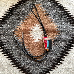 May include: A bolo tie with a black braided leather cord and silver-toned tips. The pendant is a geometric design with red, black, blue, and orange accents. The tie rests on a patterned textile.