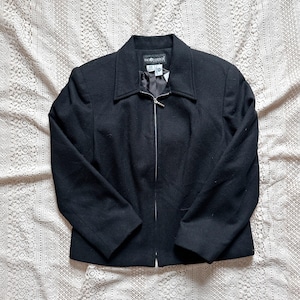 Puede incluir: Chaqueta negra con cuello y cremallera plateada. La chaqueta tiene mangas largas y un corte entallado. La chaqueta está hecha de una tela negra lisa. La chaqueta se muestra sobre una superficie de encaje blanco.