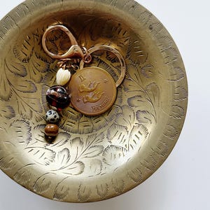 Puede incluir: Un llavero de metal dorado con un dije circular grabado con el símbolo del zodiaco Géminis y el texto GEMINI. El llavero incluye un cierre de mosquetón y una hilera de cuatro cuentas decorativas con patrones en crema, negro, marrón y blanco.