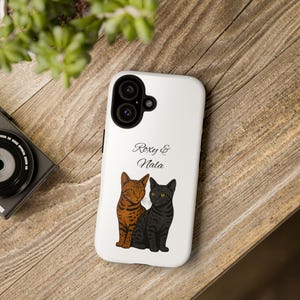 Könnte beinhalten: Eine weiße Smartphone-Hülle mit einer grafischen Illustration von zwei nebeneinander sitzenden Katzen. Eine Katze ist braun mit dunklen Streifen, die andere ist einfarbig schwarz. Der Text "Roxy & Nala" steht in schwarzer Schreibschrift darüber.