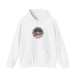 Könnte beinhalten: Ein weißer Kapuzenpullover mit einer kreisförmigen Grafik auf der Brust. Das Motiv zeigt einen Van und einen Wohnwagen auf einer Straße unter Sonne und Feuerwerk, mit dem Text "STARS, STRIPES & ROAD TRIPS" in einem Bogen und in Rot.