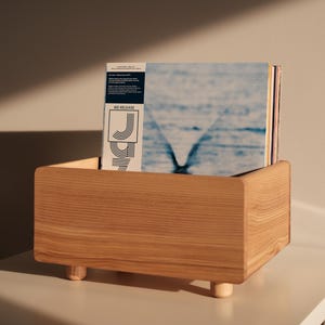 Può includere: Un portadischi in legno con finitura naturale, contenente diversi dischi in vinile. I dischi hanno una copertina blu e bianca. Il portadischi ha una forma quadrata e poggia su gambe corte e arrotondate. Il testo "WE RELEASE" è visibile.