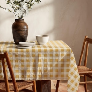 May include: A rectangular table covered with a yellow and white gingham tablecloth. On the table sits a dark brown ceramic vase filled with white flowers, a plain white ceramic bowl, and a stack of three white ceramic plates. Two wooden chairs.