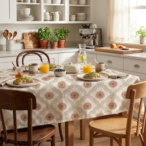 Puede incluir: Un mantel rectangular con un patrón repetitivo de soles de color terracota encerrados en contornos festoneados de color gris claro sobre una tela de color crema, extendido sobre una mesa de comedor con platos de tostadas de aguacate y vasos de zumo.