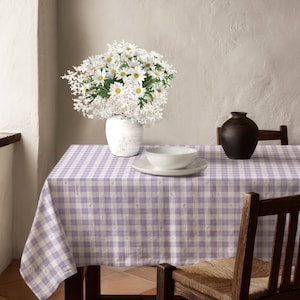 May include: A light purple and white checkered tablecloth featuring a subtle floral pattern. On the table sits a white vase filled with white daisies and baby's breath, a white bowl on a white plate, and a dark brown ceramic vase.