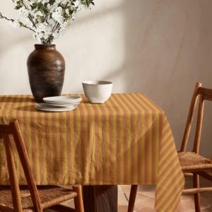 Könnte beinhalten: Eine rechteckige Tischdecke mit vertikalen Streifen in abwechselnden Senfgelb- und gedämpften Orangetönen. Der Stoff liegt auf einem Tisch, auf dem eine dunkelbraune Keramikvase mit weißen Blumen sowie eine weiße Schale stehen.