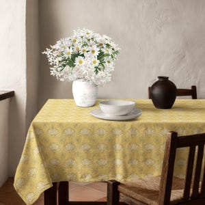 May include: A yellow tablecloth with a repeating white fan and sunburst pattern covers a table. On the table sits a white vase filled with white daisies and baby's breath, a white bowl on a white plate, and a dark brown ceramic vase.