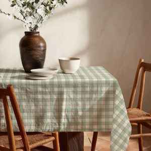 Könnte beinhalten: Eine rechteckige Tischdecke mit einem hellen salbeigrünen und weißen Vichy-Karomuster mit dünnen braunen Gitterlinien. Die Tischdecke liegt auf einem Tisch, zusammen mit einer dunkelbraunen Keramikvase, weißen Blumen und einer weißen Schale.