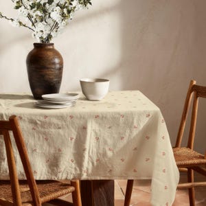 Könnte beinhalten: Eine cremefarbene Tischdecke mit einem kleinen, sich wiederholenden Blumenmuster in Rosa und Grün. Auf dem Tisch stehen eine dunkelbraune, strukturierte Vase mit weißen Blumen, drei gestapelte weiße Teller und eine weiße Schale.