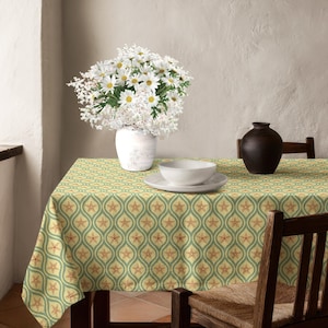 Può includere: Una tovaglia rettangolare con un motivo ripetuto di stelle marine arancioni centrate all'interno di forme a ogiva verde salvia su uno sfondo giallo pallido. Un vaso bianco riempito di margherite bianche e un vaso marrone scuro sono sul tavolo.