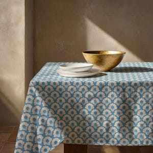 May include: A blue and white floral patterned tablecloth draped over a table. On top of the tablecloth sits a stack of two white ceramic plates and a large, textured gold-toned bowl. The pattern consists of repeating stylized flower shapes.