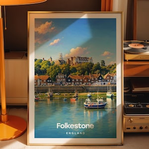 May include: A framed vertical art print featuring a coastal scene of Folkestone, England. The image displays a harbour with small boats on blue water, a row of houses, and trees under a bright sky. The text Folkestone ENGLAND is printed at the bottom.