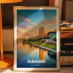 Puede incluir: Una impresión artística vertical enmarcada que presenta una colorida ilustración del horizonte de Adelaida, Australia, al atardecer. La imagen muestra un río que refleja los edificios de la ciudad y las nubes, con el texto Adelaide Australia.