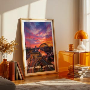 A vibrant Newcastle upon Tyne artwork rendered in high-detail illustrative style, placing the Tyne Bridge at the heart of the composition with Grey Street's Georgian sweep and Grey's Monument rising above the River Tyne in warm layered tones. Bold white NEWCASTLE UPON TYNE typography sits beneath drifting illustrated clouds. Gold frame leaning against a cream wall on parquet flooring, paired with an amber glass vase, stacked books, and an amber side table with an orange dome lamp.