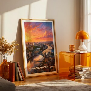 A vibrant Newcastle upon Tyne artwork rendered in high-detail illustrative style, placing the Tyne Bridge at the heart of the composition with the Millennium Bridge and warm Victorian quayside terraces rising beside the River Tyne in rich layered tones. Bold white NEWCASTLE typography sits beneath drifting illustrated clouds. Gold frame leaning against a cream wall on parquet flooring, paired with an amber glass vase, stacked books, and an amber side table with an orange dome lamp.