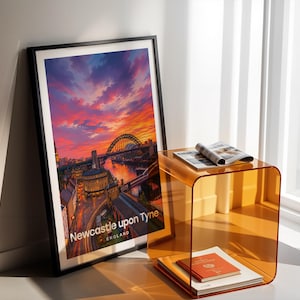 Newcastle upon Tyne cityscape poster presenting the Tyne Bridge over the River Tyne alongside the curved Georgian grandeur of Grey Street and Grey's Monument in bold illustrative colour, with crisp white NEWCASTLE UPON TYNE typography and birds circling an open illustrated sky. Black frame leaning in a white-walled corner beside an amber acrylic side table, an open magazine, a red-bound book, and warm afternoon shadows from a nearby window.