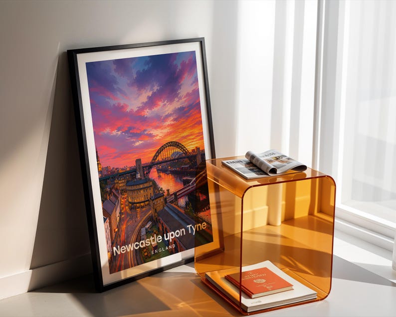 Newcastle upon Tyne cityscape poster presenting the Tyne Bridge over the River Tyne alongside the curved Georgian grandeur of Grey Street and Grey's Monument in bold illustrative colour, with crisp white NEWCASTLE UPON TYNE typography and birds circling an open illustrated sky. Black frame leaning in a white-walled corner beside an amber acrylic side table, an open magazine, a red-bound book, and warm afternoon shadows from a nearby window.