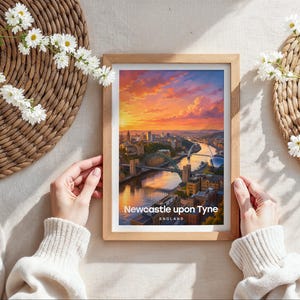 Capturing Newcastle upon Tyne in warm illustrative colour, this framed print showcases the Tyne Bridge and Millennium Bridge arching over the River Tyne with the Victorian quayside rising beyond, finished with bold white NEWCASTLE typography and birds drifting through an open illustrated sky. Oak frame laid flat on a cream linen surface with a rattan oval placemat, white daisy blooms, and hands holding the frame in soft natural daylight.