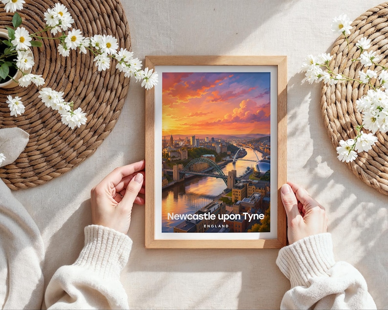 Capturing Newcastle upon Tyne in warm illustrative colour, this framed print showcases the Tyne Bridge and Millennium Bridge arching over the River Tyne with the Victorian quayside rising beyond, finished with bold white NEWCASTLE typography and birds drifting through an open illustrated sky. Oak frame laid flat on a cream linen surface with a rattan oval placemat, white daisy blooms, and hands holding the frame in soft natural daylight.