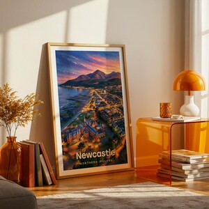 A vibrant Newcastle artwork rendered in high-detail illustrative style, placing the curved sandy beach and promenade below the dramatic rise of Slieve Donard and the sweeping Mourne Mountains beyond the rooftops. Clean white NEWCASTLE typography contrasts against a luminous illustrated sky. Displayed in a gold frame leaned on parquet flooring beside an amber glass vase and stacked books, warm amber lamp glowing nearby.