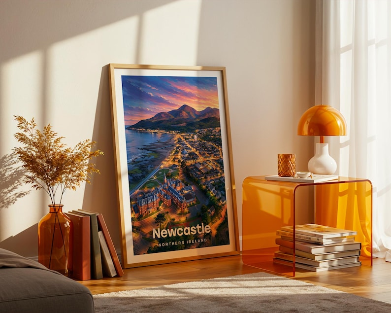 A vibrant Newcastle artwork rendered in high-detail illustrative style, placing the curved sandy beach and promenade below the dramatic rise of Slieve Donard and the sweeping Mourne Mountains beyond the rooftops. Clean white NEWCASTLE typography contrasts against a luminous illustrated sky. Displayed in a gold frame leaned on parquet flooring beside an amber glass vase and stacked books, warm amber lamp glowing nearby.