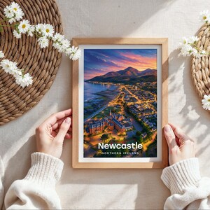 Capturing Newcastle in warm illustrative colour, this framed coastal cityscape print places the sandy beach, Slieve Donard, and the Mourne Mountains at its heart beneath drifting clouds and circling birds. Bold white NEWCASTLE typography completes the design. Shown as a flat lay on cream linen with rattan placemat, white daisy blooms, and hands holding the oak frame in soft daylight.