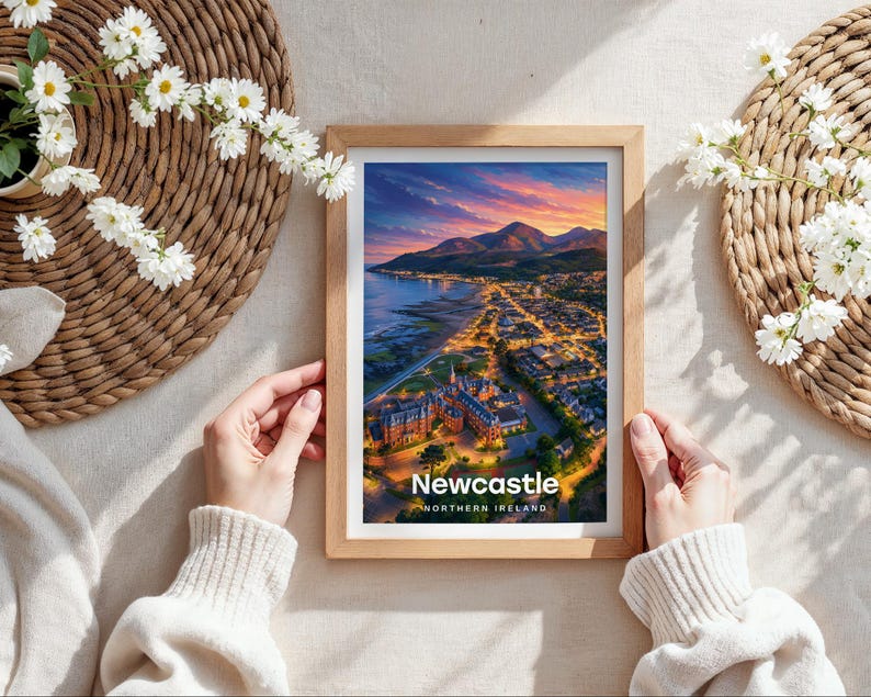 Capturing Newcastle in warm illustrative colour, this framed coastal cityscape print places the sandy beach, Slieve Donard, and the Mourne Mountains at its heart beneath drifting clouds and circling birds. Bold white NEWCASTLE typography completes the design. Shown as a flat lay on cream linen with rattan placemat, white daisy blooms, and hands holding the oak frame in soft daylight.