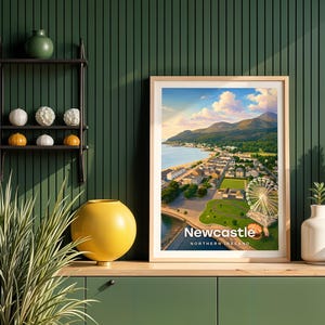 This contemporary Newcastle print celebrates the dramatic meeting of mountain and sea — Slieve Donard plunging to the sandy shore, the promenade curving below, and the Mourne ridge stretching wide across the horizon. Bold white NEWCASTLE typography sits beneath an open sky of drifting clouds and circling birds. Framed in light oak against a dark green shiplap wall with a black metal shelving unit and yellow globe vase.