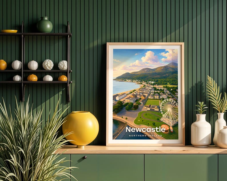 This contemporary Newcastle print celebrates the dramatic meeting of mountain and sea — Slieve Donard plunging to the sandy shore, the promenade curving below, and the Mourne ridge stretching wide across the horizon. Bold white NEWCASTLE typography sits beneath an open sky of drifting clouds and circling birds. Framed in light oak against a dark green shiplap wall with a black metal shelving unit and yellow globe vase.