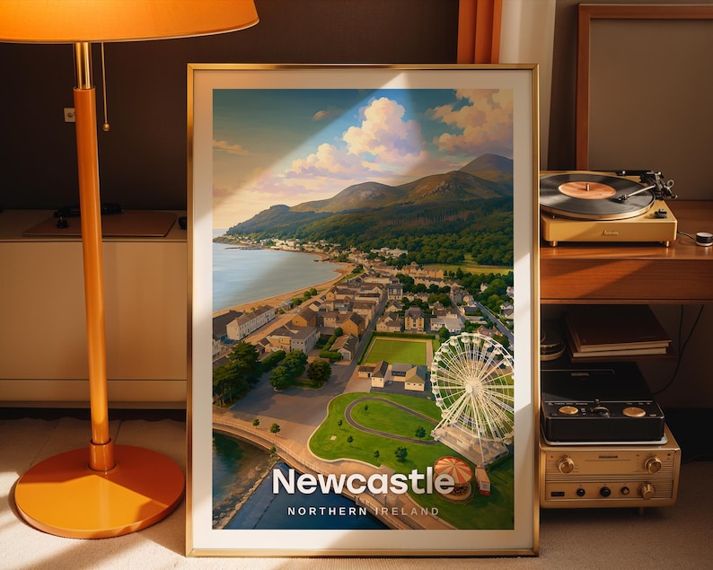 Designed as a Newcastle art print, this high-detail coastal cityscape illustration places Slieve Donard and the Mourne Mountains above the golden beach and promenade under a wide open illustrated sky. Bold white NEWCASTLE typography anchors the scene. Set in a gold frame leaned against a dark taupe wall, a warm-shaded floor lamp and vintage record player completing the vignette.