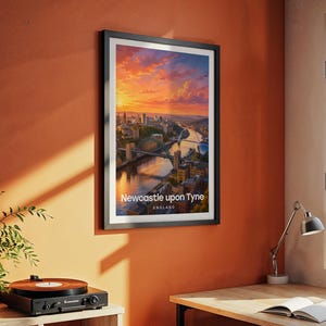 Modern Newcastle city art bringing the Tyne Bridge, Millennium Bridge, and the warm-toned Victorian quayside of the River Tyne to life in rich illustrative colour with bold white NEWCASTLE typography against a luminous illustrated sky. Black frame hung on a textured terracotta wall in a room styled with a vintage record player, yellow desk lamp, open book, green plant, and an angled metal work lamp.