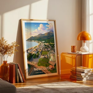 A vibrant Newcastle artwork rendered in high-detail illustrative style, with Slieve Donard's peak commanding the composition above the golden beach, the promenade, and the warm rooftops of the seaside town below the Mournes. Clean white NEWCASTLE typography contrasts against a luminous illustrated sky. Displayed in a gold frame leaned on parquet flooring beside an amber glass vase and stacked books, warm amber lamp glowing nearby.