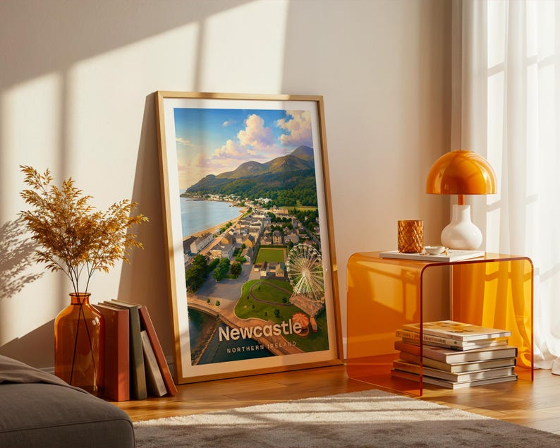 A vibrant Newcastle artwork rendered in high-detail illustrative style, with Slieve Donard's peak commanding the composition above the golden beach, the promenade, and the warm rooftops of the seaside town below the Mournes. Clean white NEWCASTLE typography contrasts against a luminous illustrated sky. Displayed in a gold frame leaned on parquet flooring beside an amber glass vase and stacked books, warm amber lamp glowing nearby.