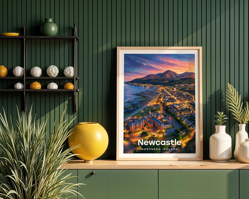 This contemporary Newcastle print celebrates the sandy promenade beach, the mighty peak of Slieve Donard climbing straight from the shore, and the rolling Mourne Mountains framing the seaside town in rich warm tones. Bold white NEWCASTLE typography sits beneath an open sky of drifting clouds and circling birds. Framed in light oak against a dark green shiplap wall with a black metal shelving unit and yellow globe vase.