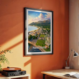 Modern Newcastle city art bringing Slieve Donard's peak, the golden sandy beach, the curved promenade, and the full sweep of the Mourne Mountains to life in warm illustrated tones with bold white NEWCASTLE typography. Framed in black and hung on a terracotta textured wall beside a vintage record player, yellow lamp, and open book in a characterful reading corner.