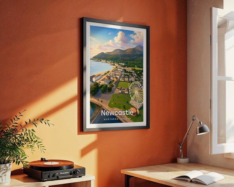 Modern Newcastle city art bringing Slieve Donard's peak, the golden sandy beach, the curved promenade, and the full sweep of the Mourne Mountains to life in warm illustrated tones with bold white NEWCASTLE typography. Framed in black and hung on a terracotta textured wall beside a vintage record player, yellow lamp, and open book in a characterful reading corner.