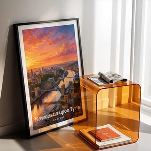 Newcastle upon Tyne cityscape poster presenting the Tyne Bridge and Millennium Bridge spanning the River Tyne above the quayside's layered Victorian buildings, in bold illustrative colour with crisp white NEWCASTLE typography and birds circling an open illustrated sky. Black frame leaning in a white-walled corner beside an amber acrylic side table, an open magazine, a red-bound book, and warm afternoon shadows from a nearby window.