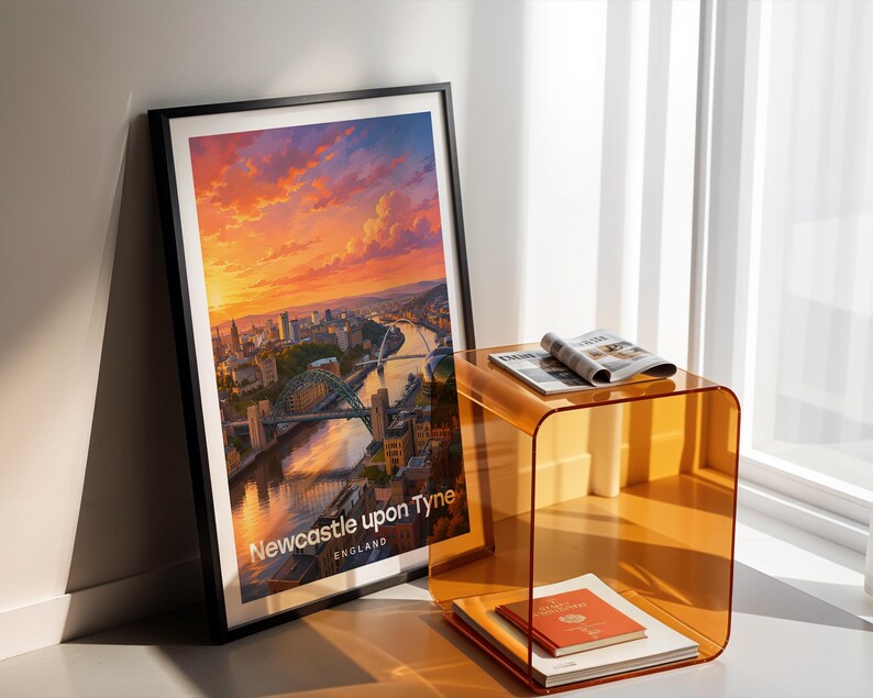 Newcastle upon Tyne cityscape poster presenting the Tyne Bridge and Millennium Bridge spanning the River Tyne above the quayside's layered Victorian buildings, in bold illustrative colour with crisp white NEWCASTLE typography and birds circling an open illustrated sky. Black frame leaning in a white-walled corner beside an amber acrylic side table, an open magazine, a red-bound book, and warm afternoon shadows from a nearby window.