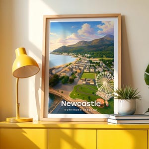 Framed Newcastle wall art print featuring a high-detail illustrated coastal scene with Slieve Donard towering above the sandy beach, the seaside promenade stretching along the shore, and the Mourne Mountains filling the horizon. Bold white NEWCASTLE typography stands out against an open illustrated sky with drifting clouds and circling birds. Displayed in a light oak frame on a cream wall beside a yellow console and warm lamp light.