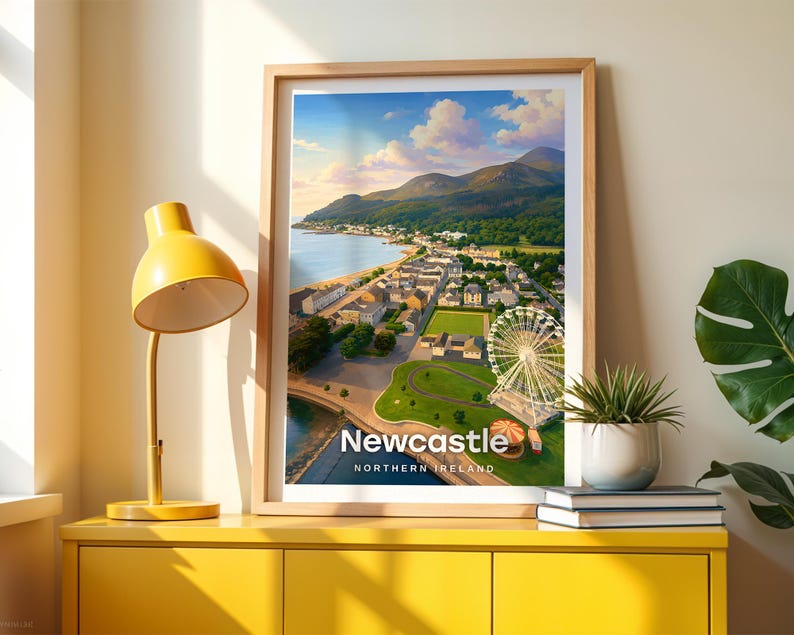 Framed Newcastle wall art print featuring a high-detail illustrated coastal scene with Slieve Donard towering above the sandy beach, the seaside promenade stretching along the shore, and the Mourne Mountains filling the horizon. Bold white NEWCASTLE typography stands out against an open illustrated sky with drifting clouds and circling birds. Displayed in a light oak frame on a cream wall beside a yellow console and warm lamp light.