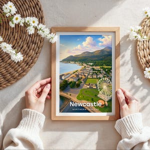Capturing Newcastle in warm illustrative colour, this framed coastal print centres Slieve Donard rising from the shore, the sandy promenade beach, and the Mourne Mountains at its heart beneath drifting clouds and circling birds. Bold white NEWCASTLE typography completes the design. Shown as a flat lay on cream linen with rattan placemat, white daisy blooms, and hands holding the oak frame in soft daylight.