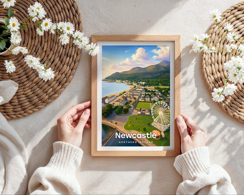 Capturing Newcastle in warm illustrative colour, this framed coastal print centres Slieve Donard rising from the shore, the sandy promenade beach, and the Mourne Mountains at its heart beneath drifting clouds and circling birds. Bold white NEWCASTLE typography completes the design. Shown as a flat lay on cream linen with rattan placemat, white daisy blooms, and hands holding the oak frame in soft daylight.
