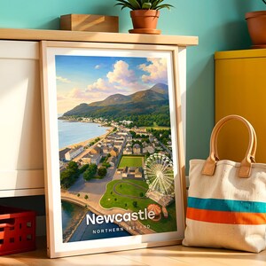 Newcastle poster capturing the Mourne Mountains sweeping down to the sandy beach, Slieve Donard dominating the skyline above the promenade, and the seaside town glowing in warm illustrative colour. Bold white NEWCASTLE typography anchors the composition beneath drifting clouds. Framed in light oak, leaned against a teal wall beside a white cabinet and terracotta planted pots.