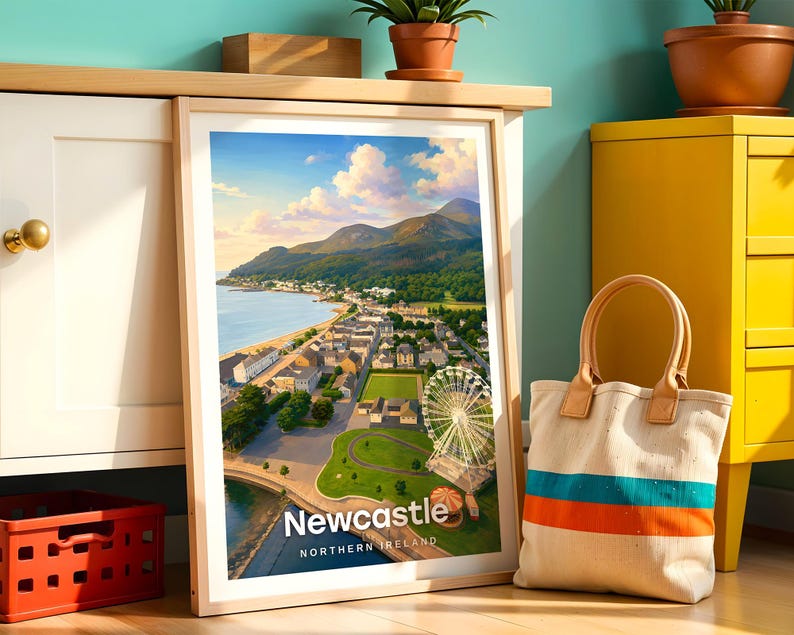 Newcastle poster capturing the Mourne Mountains sweeping down to the sandy beach, Slieve Donard dominating the skyline above the promenade, and the seaside town glowing in warm illustrative colour. Bold white NEWCASTLE typography anchors the composition beneath drifting clouds. Framed in light oak, leaned against a teal wall beside a white cabinet and terracotta planted pots.