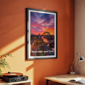 Modern Newcastle city art bringing the Tyne Bridge, the Georgian sweep of Grey Street, and Grey's Monument to life in rich warm illustrative tones with bold white NEWCASTLE UPON TYNE typography against a luminous illustrated sky. Black frame hung on a textured terracotta wall in a room styled with a vintage record player, yellow desk lamp, open book, green plant, and an angled metal work lamp.