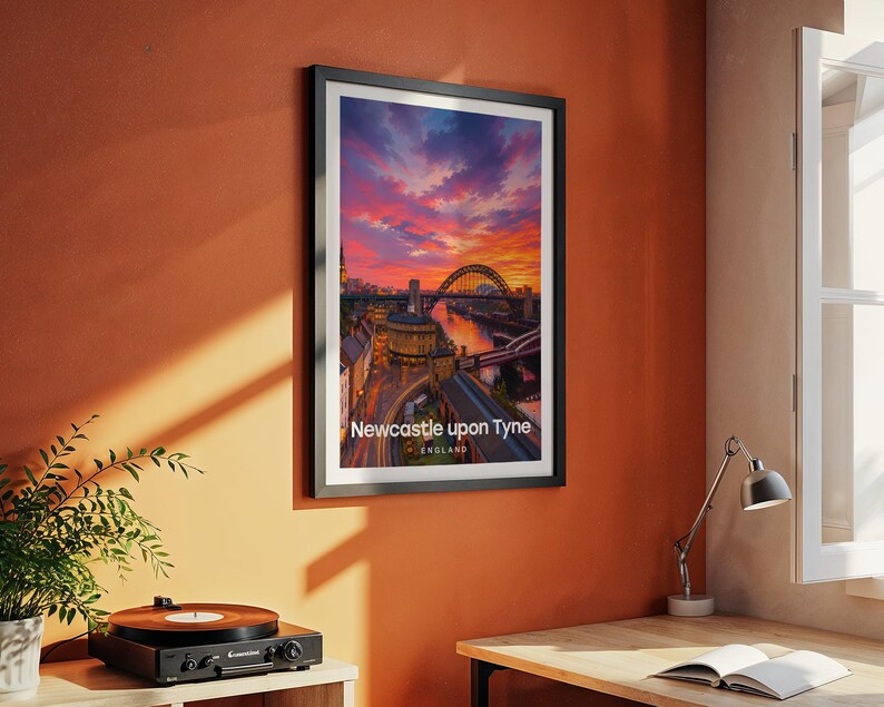 Modern Newcastle city art bringing the Tyne Bridge, the Georgian sweep of Grey Street, and Grey's Monument to life in rich warm illustrative tones with bold white NEWCASTLE UPON TYNE typography against a luminous illustrated sky. Black frame hung on a textured terracotta wall in a room styled with a vintage record player, yellow desk lamp, open book, green plant, and an angled metal work lamp.