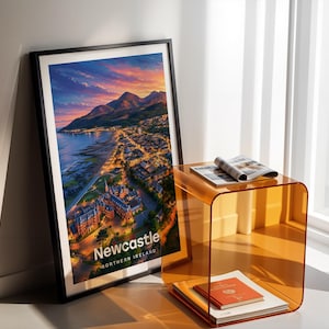Newcastle cityscape poster presenting the sandy beach, the peak of Slieve Donard, and the Mourne Mountains above the seaside town in bold illustrative colour with clean white NEWCASTLE typography overhead. An open sky of drifting clouds and circling birds fills the upper composition. Framed in black, leaned in a white-walled corner beside an amber acrylic side table and an open magazine in warm shadow light.
